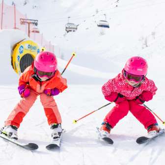 Deux enfants avec des casques roses font du ski en riant sur la piste de Malbun par un temps hivernal ensoleillé.