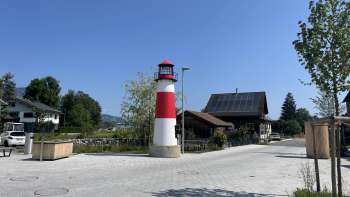 Petit phare rouge et blanc Moby sur une place de Ruggell par temps ensoleillé, avec des bâtiments traditionnels en arrière-plan.
