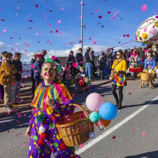 Clown déguisé avec des confettis et des ballons lors du défilé de carnaval, spectateurs au bord de la route.