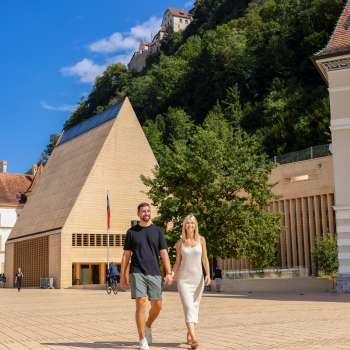 Un couple se promène sur la place ensoleillée du gouvernement à Vaduz, avec vue sur le bâtiment moderne du Landtag et le château de Vaduz en arrière-plan.