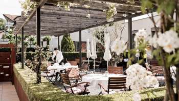 Une terrasse élégante avec des tables blanches, couvertes de serviettes et de verres, sous un pavillon en bois entouré d'une verdure luxuriante.