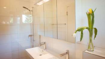 Salle de bain moderne au carrelage clair dans la maison d'hôtes près du camping Mittagsspitze avec double lavabo, miroir et fleurs fraîches.