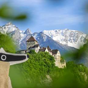 Vue sur le château de Vaduz avec la mascotte du Detektiv-Trail au premier plan et les Alpes enneigées en arrière-plan.