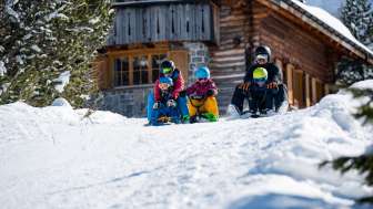 Faire de la luge avec les enfants à Malbun Deux adultes et trois enfants assis sur des luges entament ensemble une descente devant un chalet de montagne enneigé à Malbun.