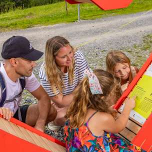Une famille avec des enfants lit avec attention le panneau d'information sur le parcours du cœur alpin à Malbun, un sentier de randonnée interactif dans les montagnes du Liechtenstein.