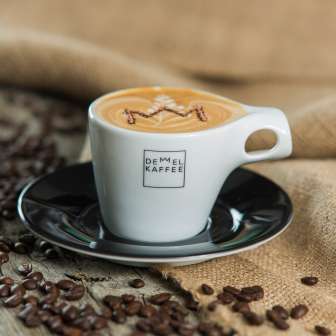Cappuccino avec couronne dans la mousse dans une tasse de Demmel Kaffee, entouré de grains de café sur du bois et un sac en jute - Le plaisir au Liechtenstein