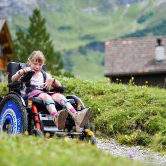 Jeune fille en fauteuil roulant électrique sur un sentier de randonnée à Malbun avec un décor de montagnes vertes en arrière-plan.