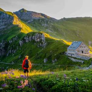 Un randonneur regarde le refuge Pfälzerhütte au Liechtenstein, entouré d'un paysage de montagnes verdoyantes et d'un panorama alpin au crépuscule.