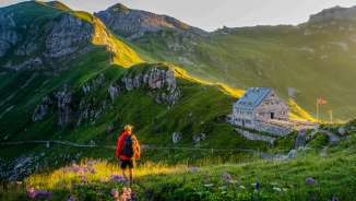 Un randonneur regarde le refuge Pfälzer au Liechtenstein, entouré d'un paysage de montagnes verdoyantes et d'un panorama alpin au crépuscule.