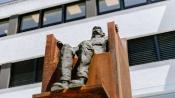 Sculpture "Hochsitz" de Robert Indermaur : personnage assis en hauteur sur une chaise en acier couleur rouille devant une façade moderne.