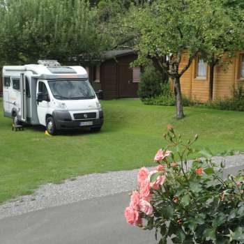 Aire de camping-car sur le camping Mittagsspitze avec une pelouse verte et des cabanes en bois entourées.