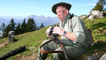 Un chasseur avec des jumelles est assis dans une prairie avec un décor de montagnes alpines.