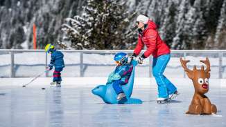 Une mère pousse son enfant sur un pingouin auxiliaire de patinage sur la patinoire de Malbun, avec en arrière-plan un enfant avec une crosse de hockey sur glace.