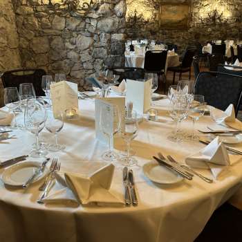 Table ronde élégamment dressée avec nappes, serviettes et menus blancs devant un mur de pierre.