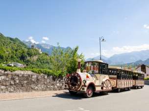 Citytrain traverse Vaduz, avec le château de Vaduz et les Alpes en arrière-plan, sous un soleil radieux.