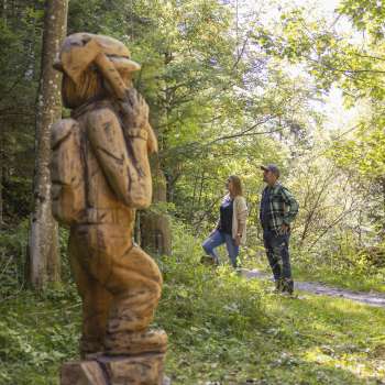 Deux randonneurs découvrent un personnage sculpté sur le « sentier des légendes walsers » au milieu d'une forêt baignée de lumière.