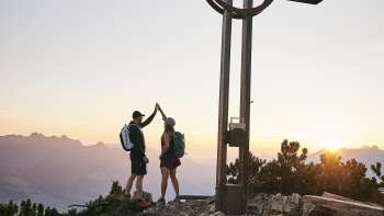 Deux randonneurs se font un high-five à la croix du sommet près de l'Alpspitz au lever du soleil avec un panorama de montagnes en arrière-plan.