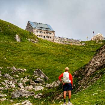 Un randonneur avec un sac à dos rouge regarde le refuge Pfälzer au milieu d'un paysage de montagne verdoyant.