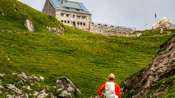 Un randonneur avec un sac à dos rouge regarde le refuge Pfälzerhütte au milieu d'un paysage de montagne verdoyant.