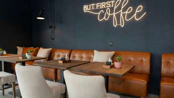 Un café moderne avec des canapés en cuir, des fauteuils blancs et des tables en bois. Sur le mur bleu foncé, l'inscription "BUT FIRST coffee" brille en néon clair. Un café moderne avec des canapés en cuir, des fauteuils blancs et des tables en bois. Sur le mur bleu foncé, l'inscription "BUT FIRST coffee" brille en néon clair.