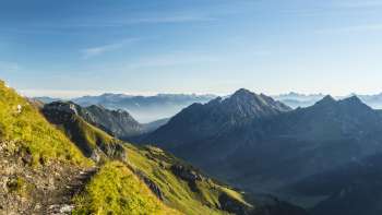 Sentier alpin avec vue sur un paysage de montagnes verdoyantes et des sommets escarpés au soleil du matin
