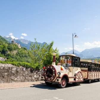 Citytrain traverse Vaduz, avec le château de Vaduz et les Alpes en arrière-plan, sous un soleil radieux.