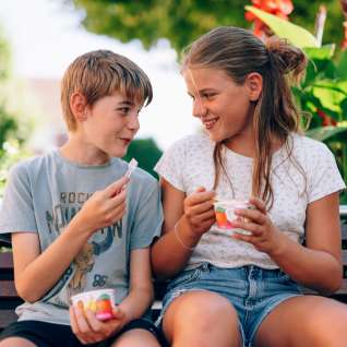 Deux enfants sont assis sur un banc dans un parc du Liechtenstein et mangent une glace - des visages souriants, des fleurs et une ambiance estivale dans la verdure.