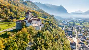 Vue sur le château de Vaduz avec la forêt aux couleurs de l'automne et la ville de Vaduz dans la vallée.