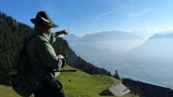 Un homme en tenue traditionnelle montre la vallée du Rhin depuis une prairie de montagne.