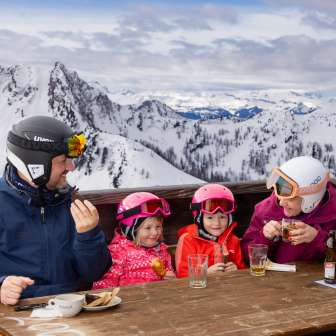 La famille profite d'une pause sur une terrasse ensoleillée de Malbun avec vue sur les montagnes enneigées.