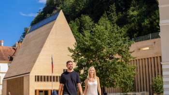 Un couple se promène sur la place ensoleillée du gouvernement à Vaduz, avec vue sur le bâtiment moderne du Landtag et le château de Vaduz en arrière-plan.