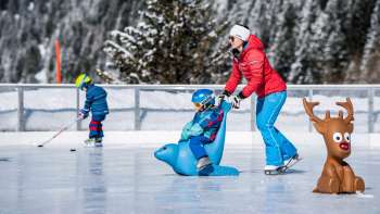 Famille sur la patinoire du Schlucher-Treff Malbun, aidée par un support de patinage en forme d'animal bleu.