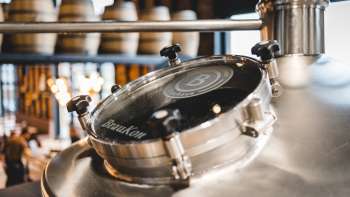Gros plan d'un hublot rond sur une cuve de brassage brillante avec vue sur la salle de brassage en arrière-plan.