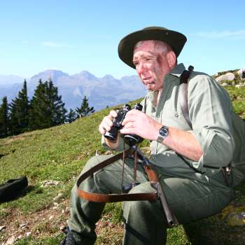 Un chasseur avec des jumelles est assis dans une prairie avec un décor de montagnes alpines.