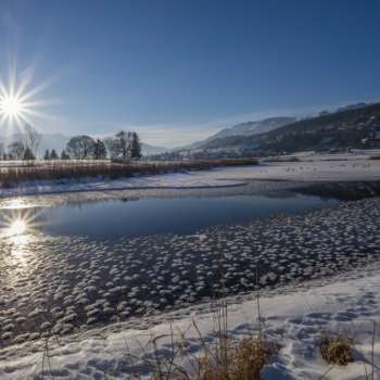 Lever de soleil sur le lac Egelsee à Mauren en hiver avec une surface de glace scintillante, de la neige et un décor de montagne ensoleillé.