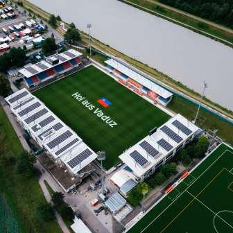 Vue aérienne du stade Rheinpark à Vaduz avec l'inscription « Hoi aus Vaduz » sur le terrain et le Rhin en arrière-plan