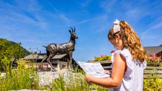 Une fillette avec une carte devant la statue de chamois à Malbun - une exploration adaptée aux enfants dans une station de vacances familiale au Liechtenstein.