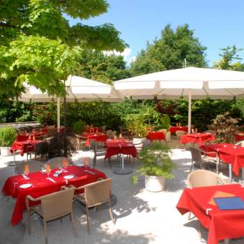 Vue extérieure de la terrasse avec des nappes rouges, entourée d'arbres et de parasols.