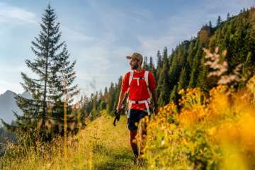 Randonneur en t-shirt rouge sur un chemin de montagne ensoleillé près de Malbun, entouré de fleurs de prairie, de forêts de conifères et d'un décor alpin au Liechtenstein.