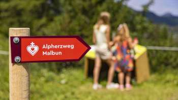 Panneau indicateur du sentier du minerai d'alpage de Malbun avec des enfants devant l'une des stations en arrière-plan