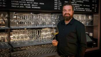 Sympathique barman du WY Weinbar avec un verre à vin devant une étagère stylée remplie de verres et de vins.