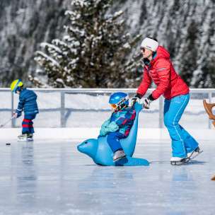 Une mère pousse son enfant sur un pingouin auxiliaire de patinage sur la patinoire de Malbun, avec en arrière-plan un enfant avec une crosse de hockey sur glace.