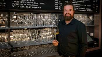 Sympathique barman du WY Weinbar avec un verre à vin devant une étagère stylée remplie de verres et de vins.