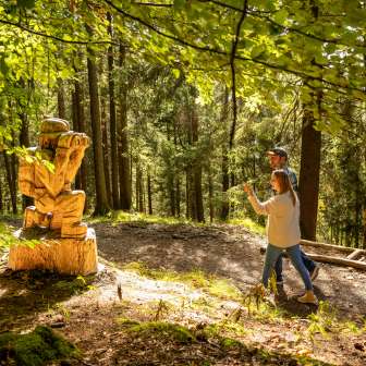 Deux visiteurs découvrent une figure en bois sculptée le long du WalserSagenWeg à Triesenberg, Liechtenstein - sentier de randonnée thématique mystique dans la forêt