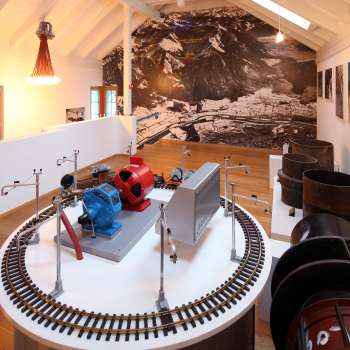 Modèle d'une turbine sur une plateforme d'exposition au musée Lawena, avec des photographies historiques en noir et blanc en arrière-plan.