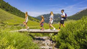 Une famille traverse un petit pont en bois lors d'une randonnée à travers le paysage alpin de Malbun, au Liechtenstein, sous un soleil radieux.