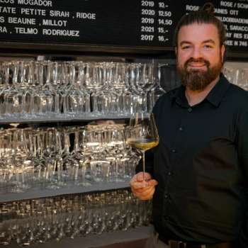 Sympathique barman du WY Weinbar avec un verre à vin devant une étagère stylée remplie de verres et de vins.