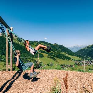 Deux enfants se balancent sur une aire de jeux de montagne avec vue sur le panorama estival de Malbun au Liechtenstein.