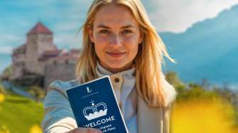 Welcome Passeport découvertes - Découvrir Vaduz & ses environs Femme tenant le WELCOME Passeport découvertes devant le château de Vaduz dans un champ en fleurs