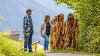Couple regardant des figures en bois sculptées sur le sentier des légendes walser à Triesenberg, avec vue sur la vallée et les montagnes en arrière-plan.